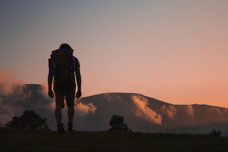 Backpack Journey - silhouette of man walking along field leading to mountain